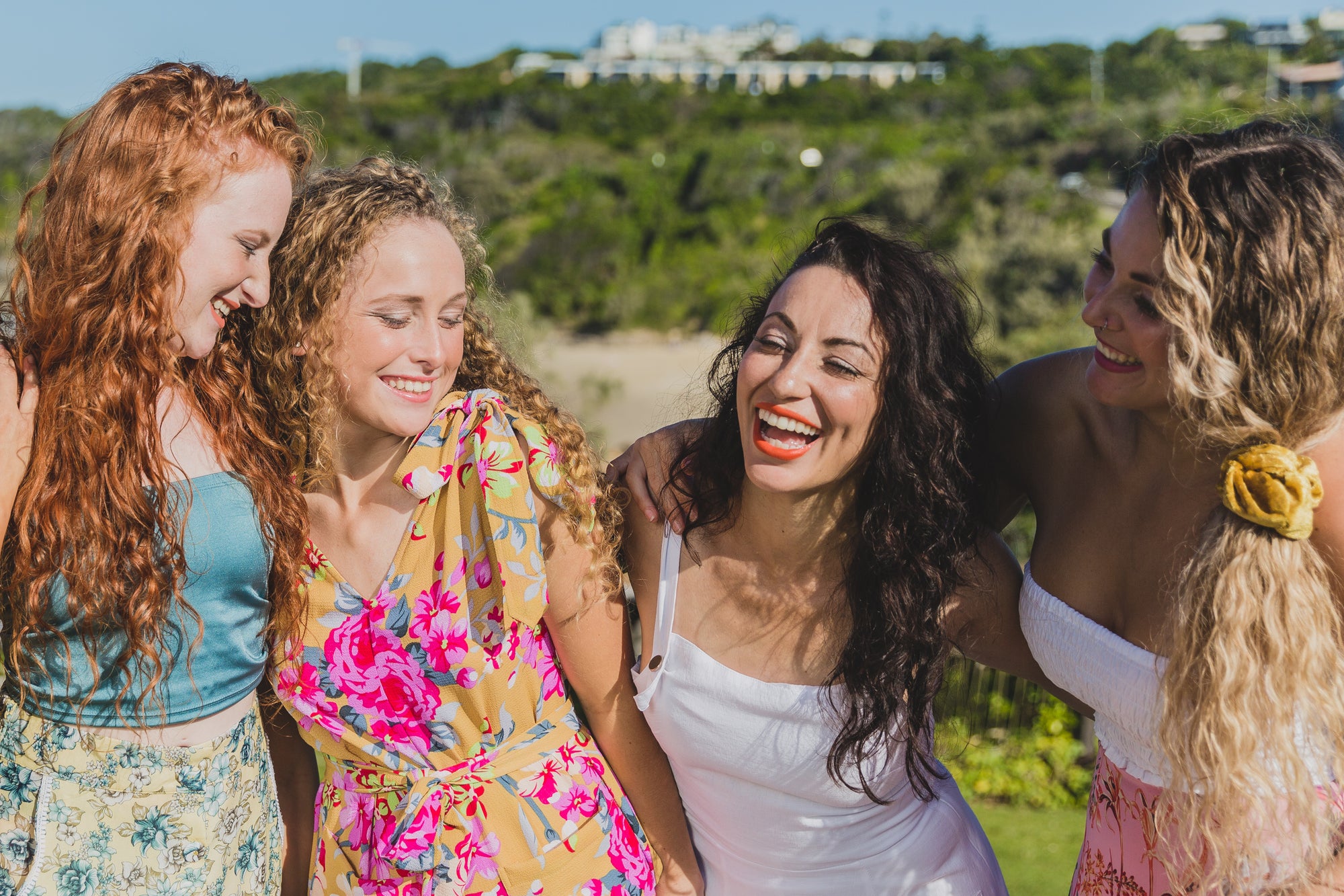 An image of 4 women with curly hair wearing bright clothing and smiling and laughing in the sunshine with their arms linked around each other.