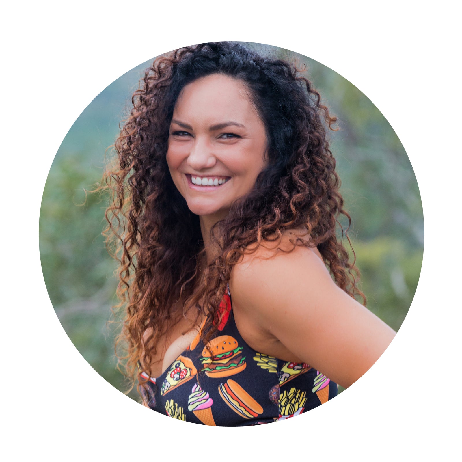 A circluar image of a women with curly brown hair, standing with one arm on her hip and her body is facing to the left and her face is directly smiling to the camera 